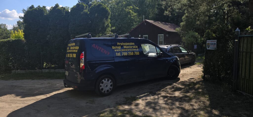 Granatowy van firmy ANTENY z napisem "Profesjonalne Usługi Montaż anten i usługi IT Pruszk&oacute;w" zaparkowany na podjeździe przed domem w słoneczny dzień.